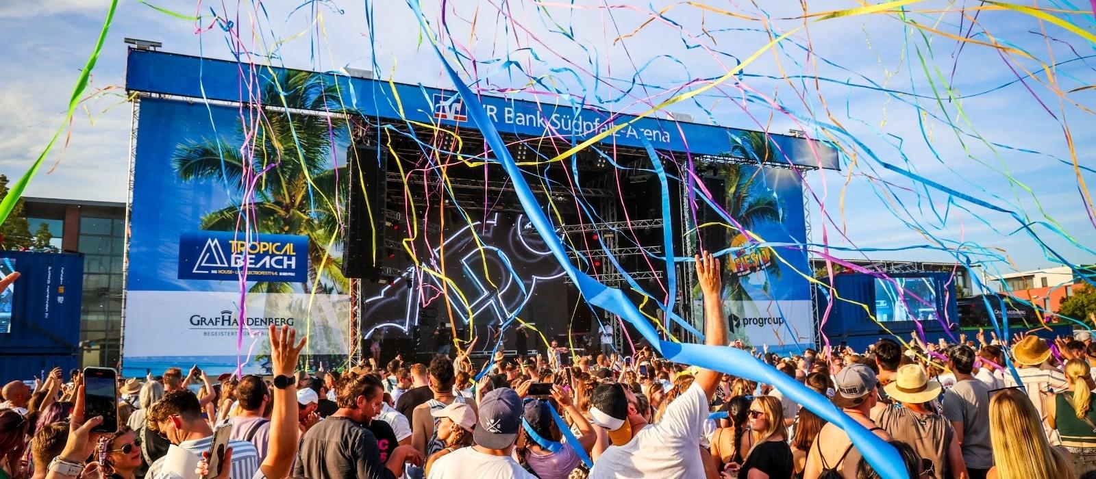 Bühne des Tropical Beach Festivals mit Palmen und Luftschlangen über der feiernden Menge